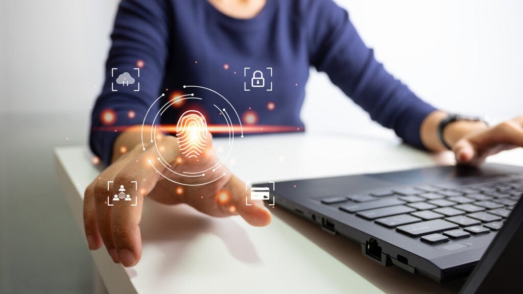 A person working on a laptop stretching out the right index finger to leave a fingerprint in the air. The symbols surrounding the finger stand for cybersecurity and compliance.