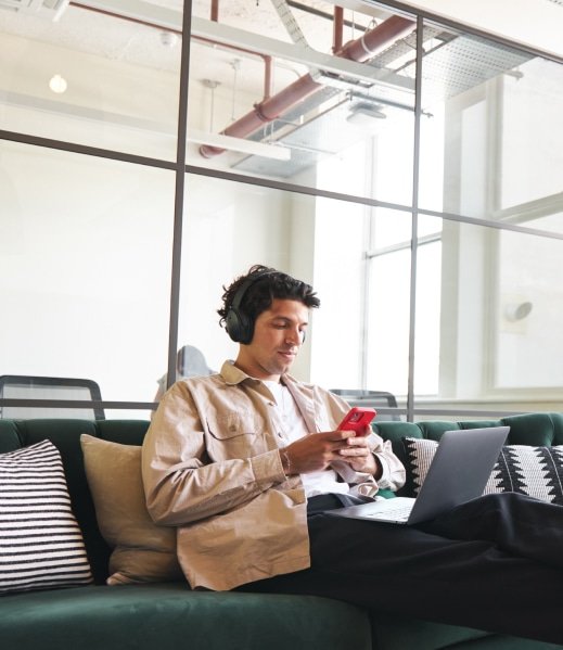 A man sitting on a sofa. He has his laptop on his lap and is looking at the phone in his hands.