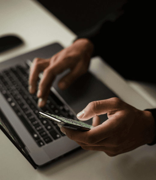 Hands of a person that is typing at a laptop with one hand and also on a mobile screen with the other